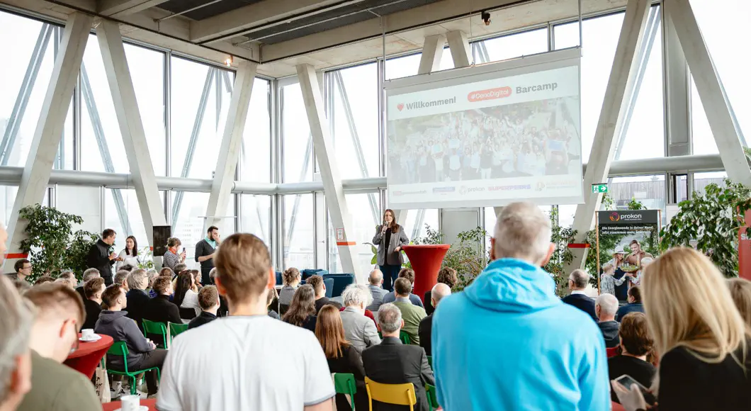 Großer heller Saal mit Bühne und Leinwand. Eine Frau spricht zu den Menschen im Saal.